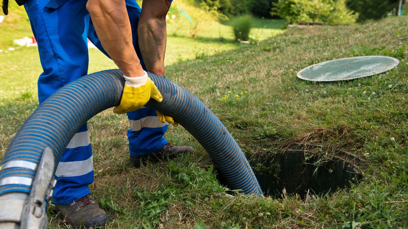 A service technician feeding a large vacuum hose into an open septic tank access port in a lawn during a routine pump out