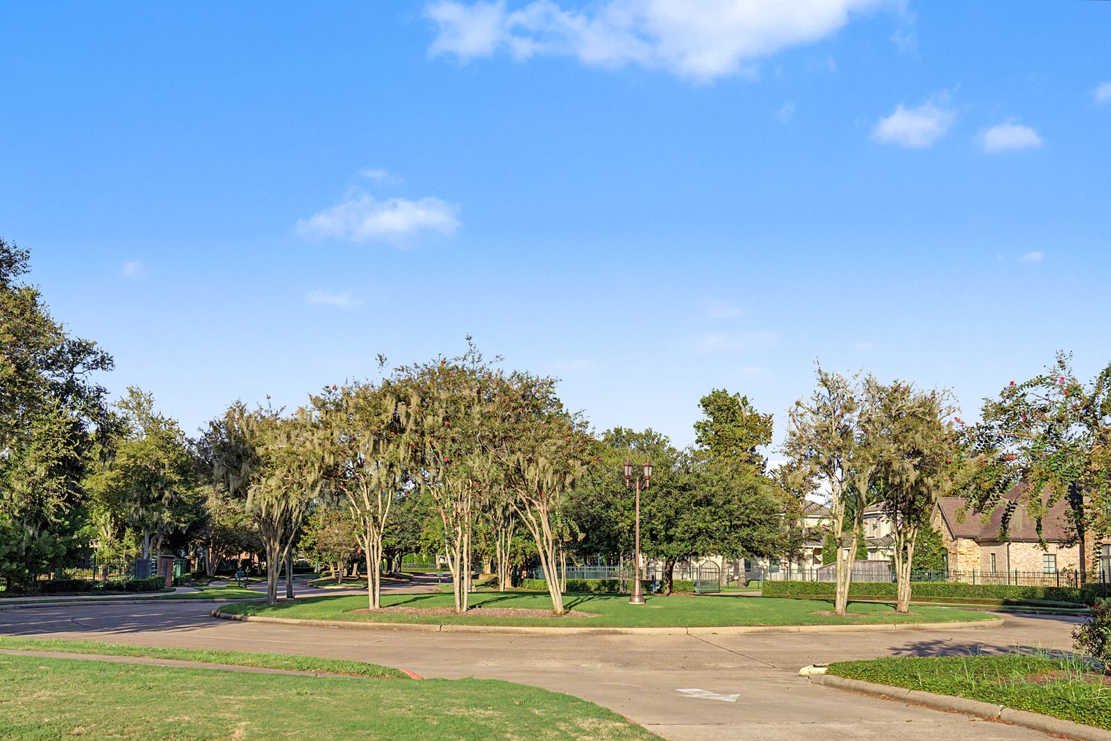A tree lined street at the entrance to Sienna Golf Club, with crepe myrtles, moss draped oaks, a gated drive and brick and stone homes beyond