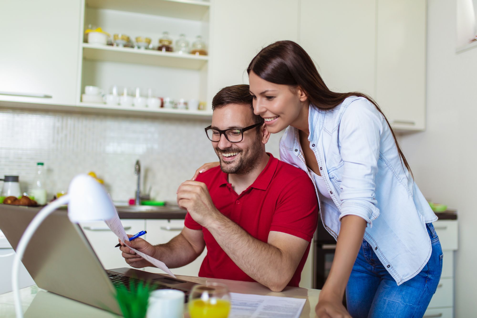Smiling couple at a kitchen table looking at a laptop, the man in glasses holding a printed document and a pen